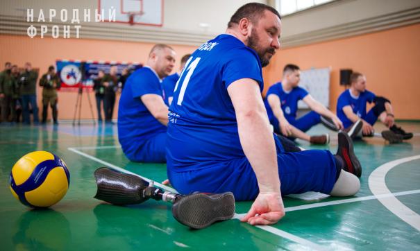 Спортсмен в форме без ноги сидит в зале, рядом волейбольный мяч и отстегнутый протез ноги, на фоне другие члены команды
