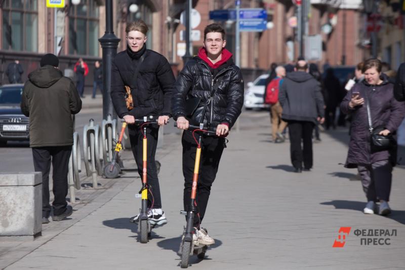 Двое молодых мужчин на электросамокатах едут по весенней улице без снега в демисезонной одежде