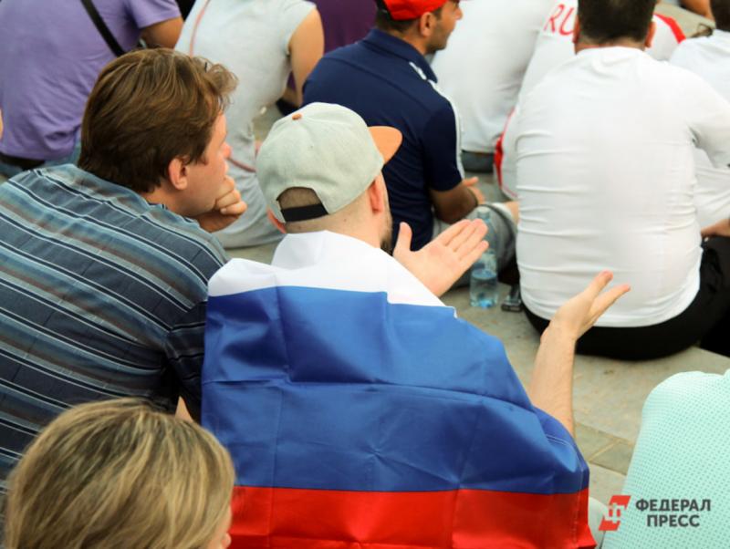 Двое болельщиков сидят спиной на трибуне на футбольном матче, один завернут в флаг России
