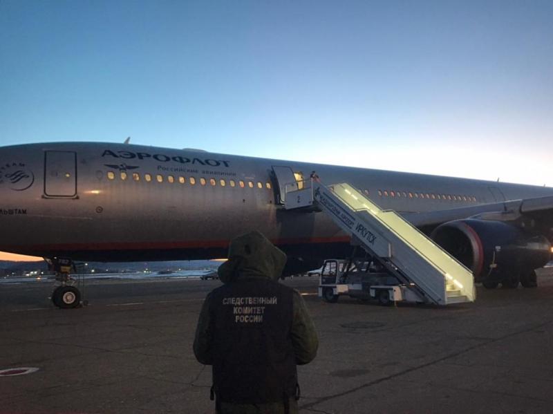 Следователь СК на транспорте стоит спиной на фоне самолета, стоящего в аэропорту с выдвинутым трапом с надписью Иркутск