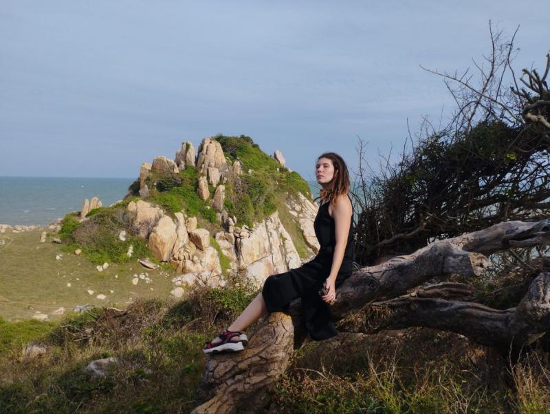 Девушка с дредами в черном платье сидит на скальном уступе, на фоне гора и море