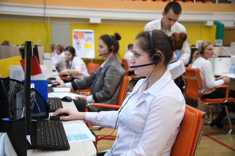 Сотрудница колл-центра в белой блузке сидит в офисе перед компьютером в наушниках с микрофоном, на фоне другие сотрудники